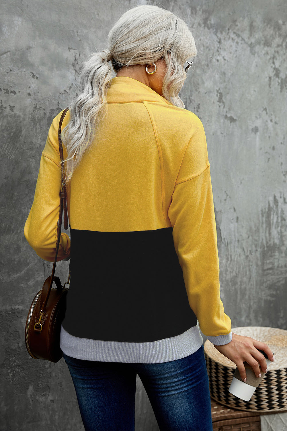 A person with long, light hair in a ponytail, wearing a Black Zipped Colorblock Sweatshirt with Pockets, blue jeans, and glasses, is holding a white cup and a brown handbag, shown from behind against a gray wall.