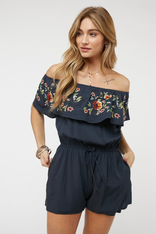 Wearing the stylish Floral Embroidered Off Shoulder Romper, a woman poses gracefully, showcasing its ruffle detail and making it an ideal addition to any summer wardrobe.