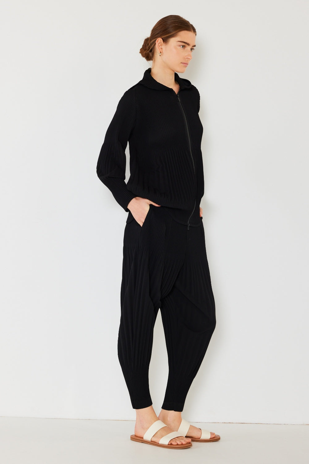 A person stands against a plain background, wearing a matching dark green hooded top and Marina West Swim Pleated Unisex Aladdin Plisse Pants with a ribbed texture, complemented by white sandals.