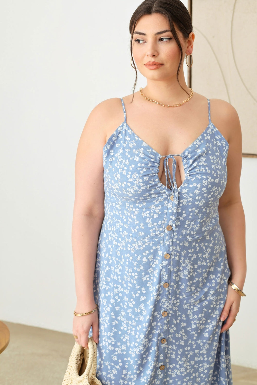 A woman stands indoors wearing the Zenobia Plus Size Cutout Floral Spaghetti Strap Dress in light blue, holding a small woven handbag. She is accessorized with earrings, bangles, and beige platform sandals.