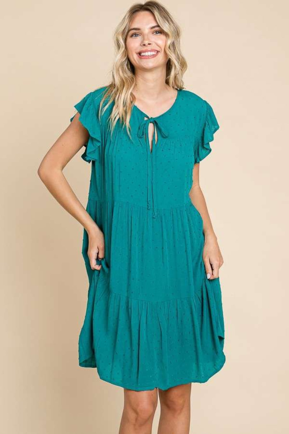 A woman stands against a beige background wearing the Culture Code Ruffle Cap Sleeve Tiered Dress, which is teal and flowing with short ruffled sleeves and a tie at the neckline. She is smiling with one hand in her pocket, showcasing the lightweight fabric.