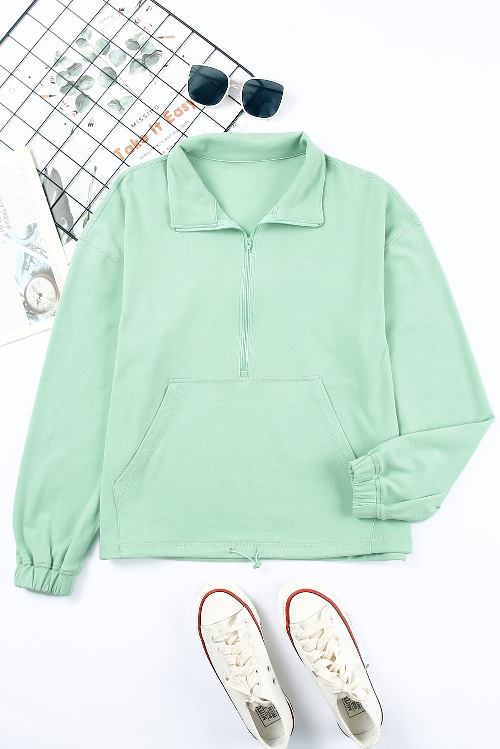 A person with wavy hair stands with their back to the camera, wearing a Green Zip Front Pocketed Pullover Sweatshirt and blue jeans, holding a small brown bag in their left hand.