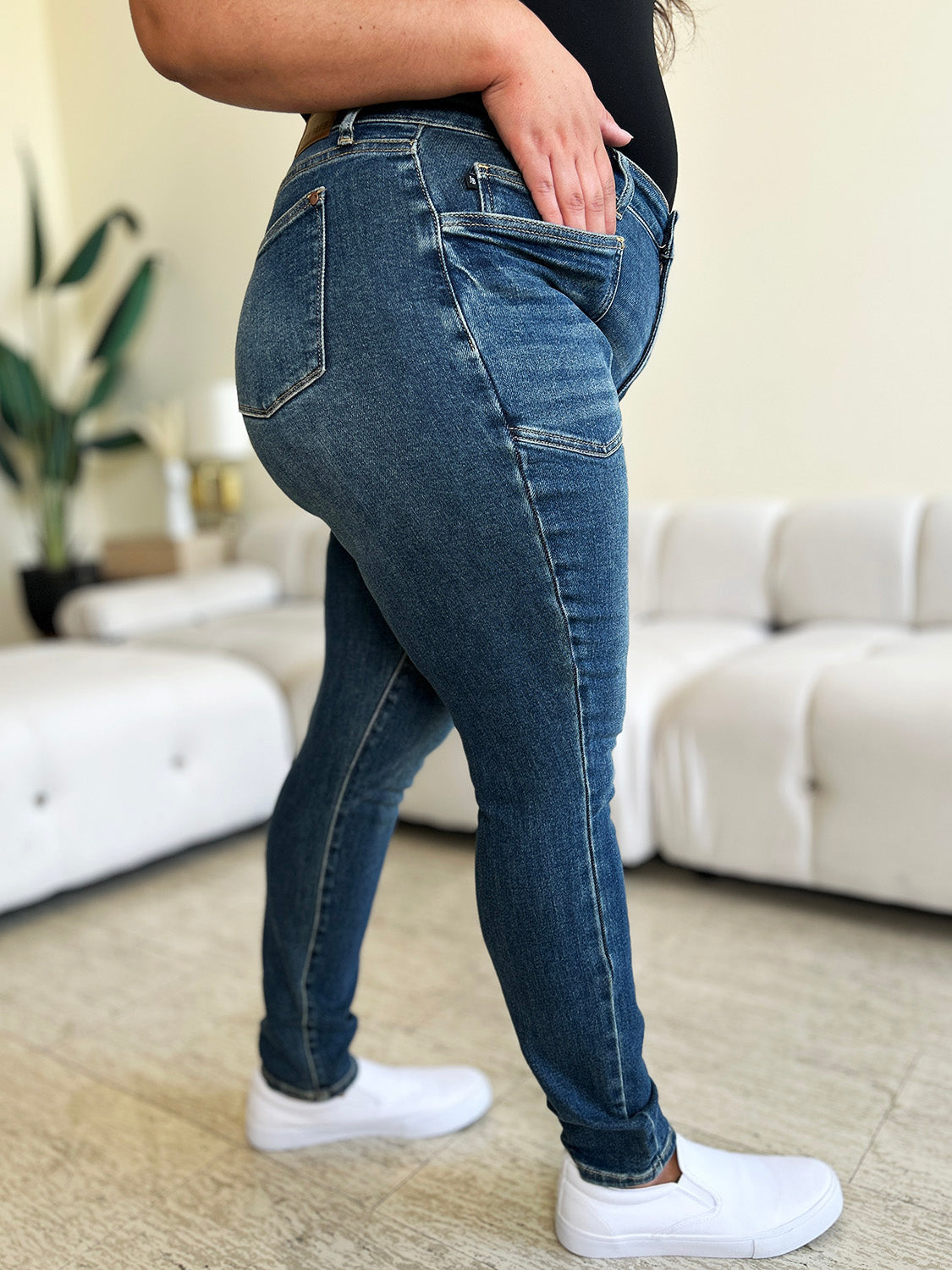 A person wearing Judy Blue Full Size High Waist Skinny Jeans made of stretchy denim fabric stands in a room with light-colored furniture and wooden flooring, paired perfectly with white slip-on shoes.