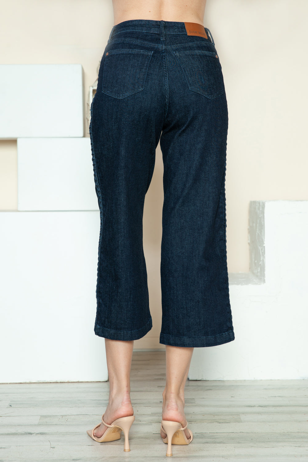 Person wearing Judy Blue Full Size Side Seam Braid Detail Crop Wide Leg Jeans and beige strappy heels, standing on a wooden floor with a white background.