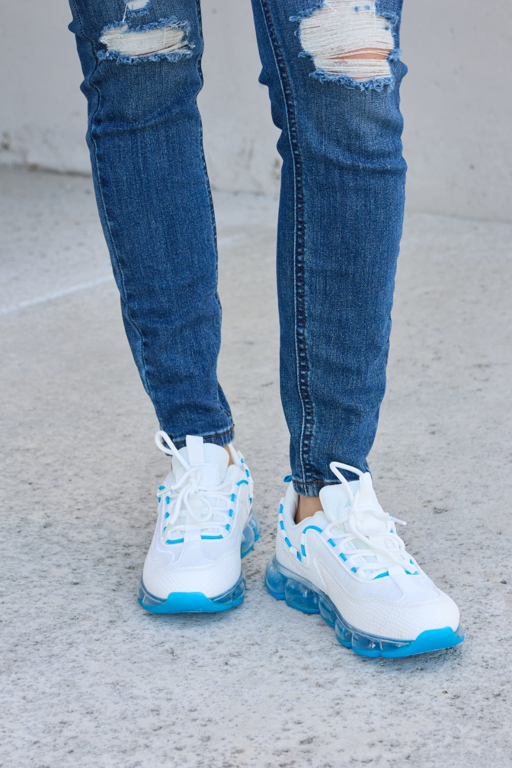 A person wearing ripped jeans and stylish Forever Link Lace-Up Air-Cushioned Athletic Shoes in white with blue accents stands confidently on a concrete surface.