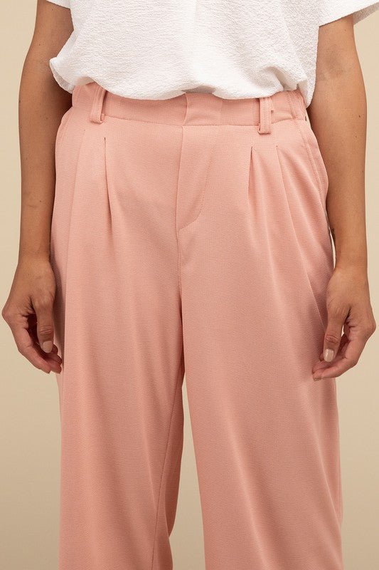 A woman stands against a beige background, highlighting her chic ensemble with a tailored fit. She wears a white short-sleeve shirt paired with versatile pink Waffle Trouser Pants, hands casually in pockets, complemented by stylish strappy sandals.