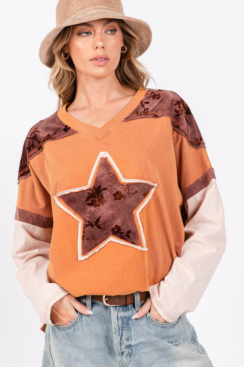 A person wearing a beige hat, a SAGE + FIG Star Patch Long Sleeve Color Block T-Shirt in orange, and light blue jeans stands against a plain background.