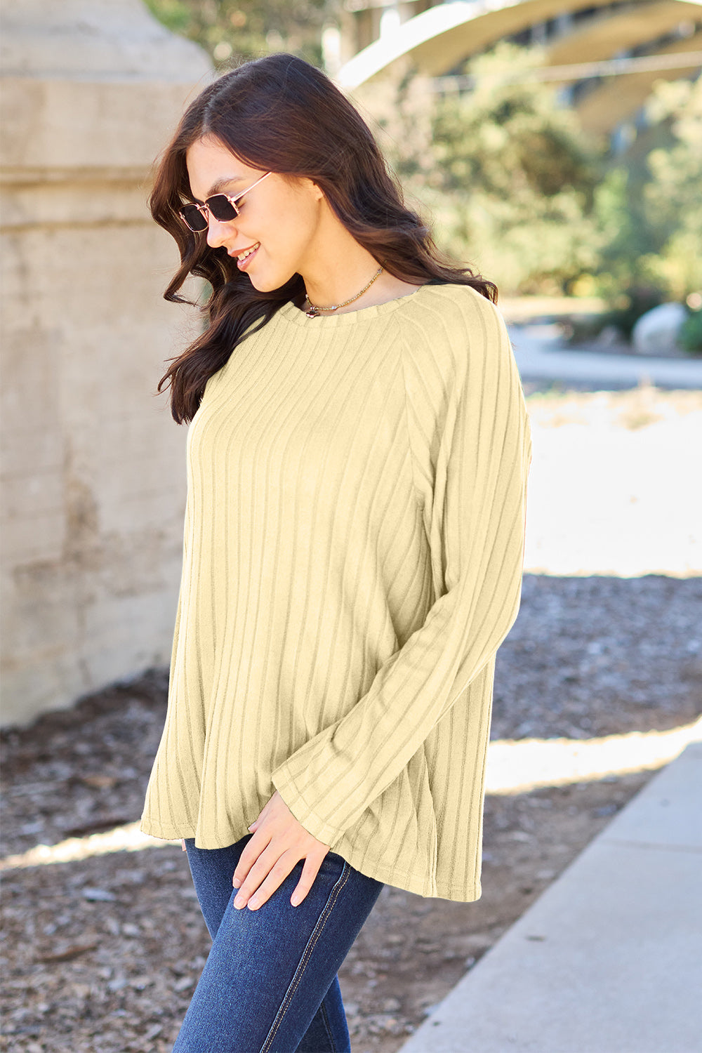 A woman with long brown hair wearing sunglasses and a Basic Bae full-size, slightly stretchy ribbed round neck long sleeve knit top in red, paired with blue jeans, is standing outdoors.