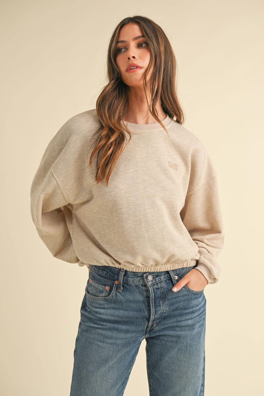 A woman with long brown hair wears the Mable Round Neck Letter Embroidery Crop Sweatshirt in beige, paired with blue jeans, standing confidently with one hand in her pocket against a plain background, perfectly capturing effortless street style.