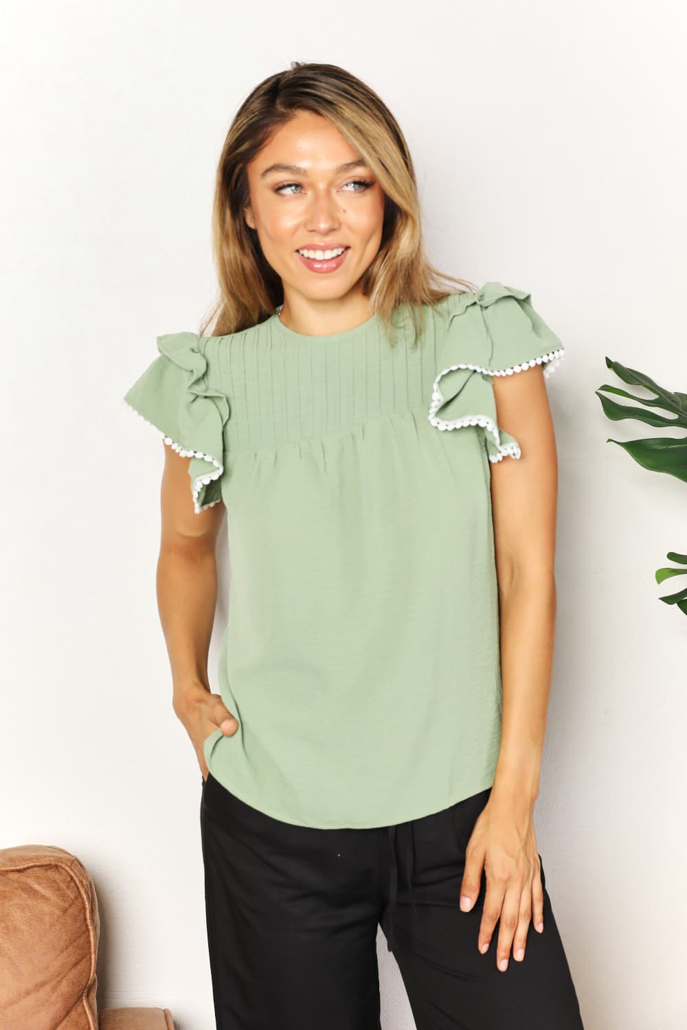 A woman stands and smiles, wearing the Perfee Pleated Detail Flutter Sleeve Blouse in light green paired with black pants.