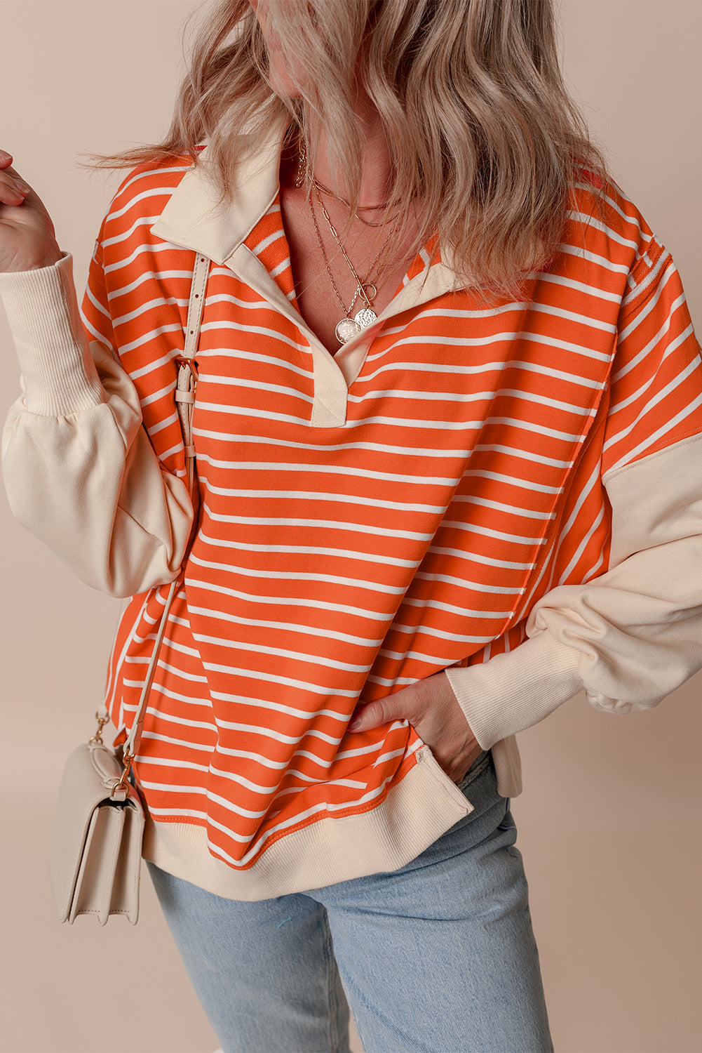 A person with wavy hair is seen from the back, wearing an Orange Stripe Color Block Loose Fit Collared Drop Shoulder Sweatshirt, light blue jeans, and carrying a small beige bag.