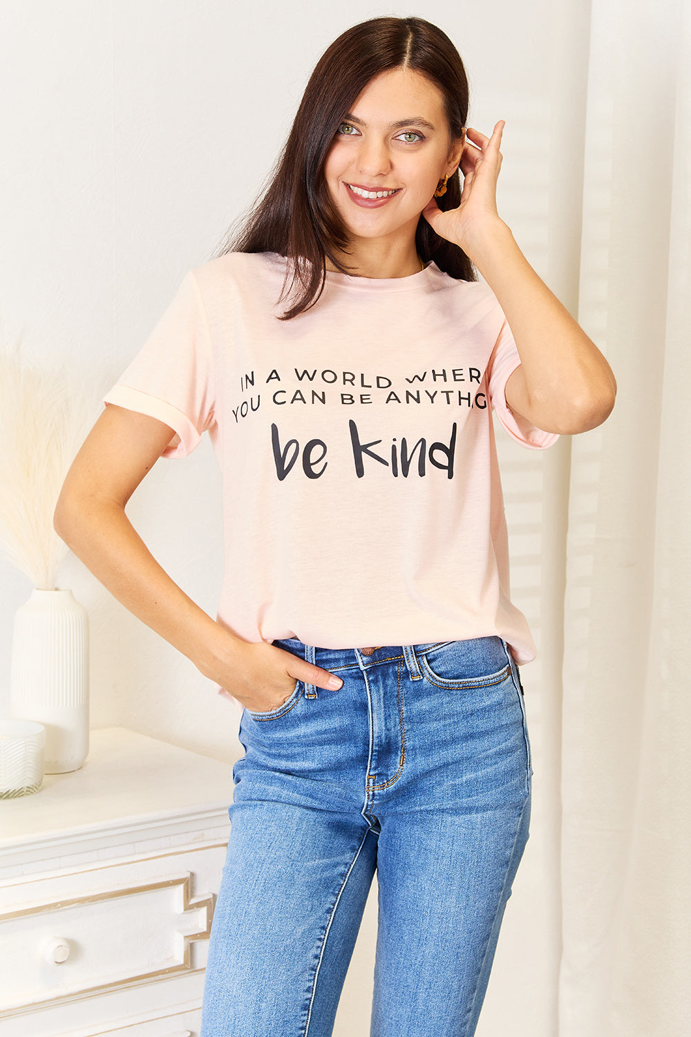 A person wears a Simply Love Slogan Graphic Cuffed T-Shirt in light pink, adorned with the inspiring message, "In a world where you can be anything, be kind," and featuring stylish cuffed sleeves.