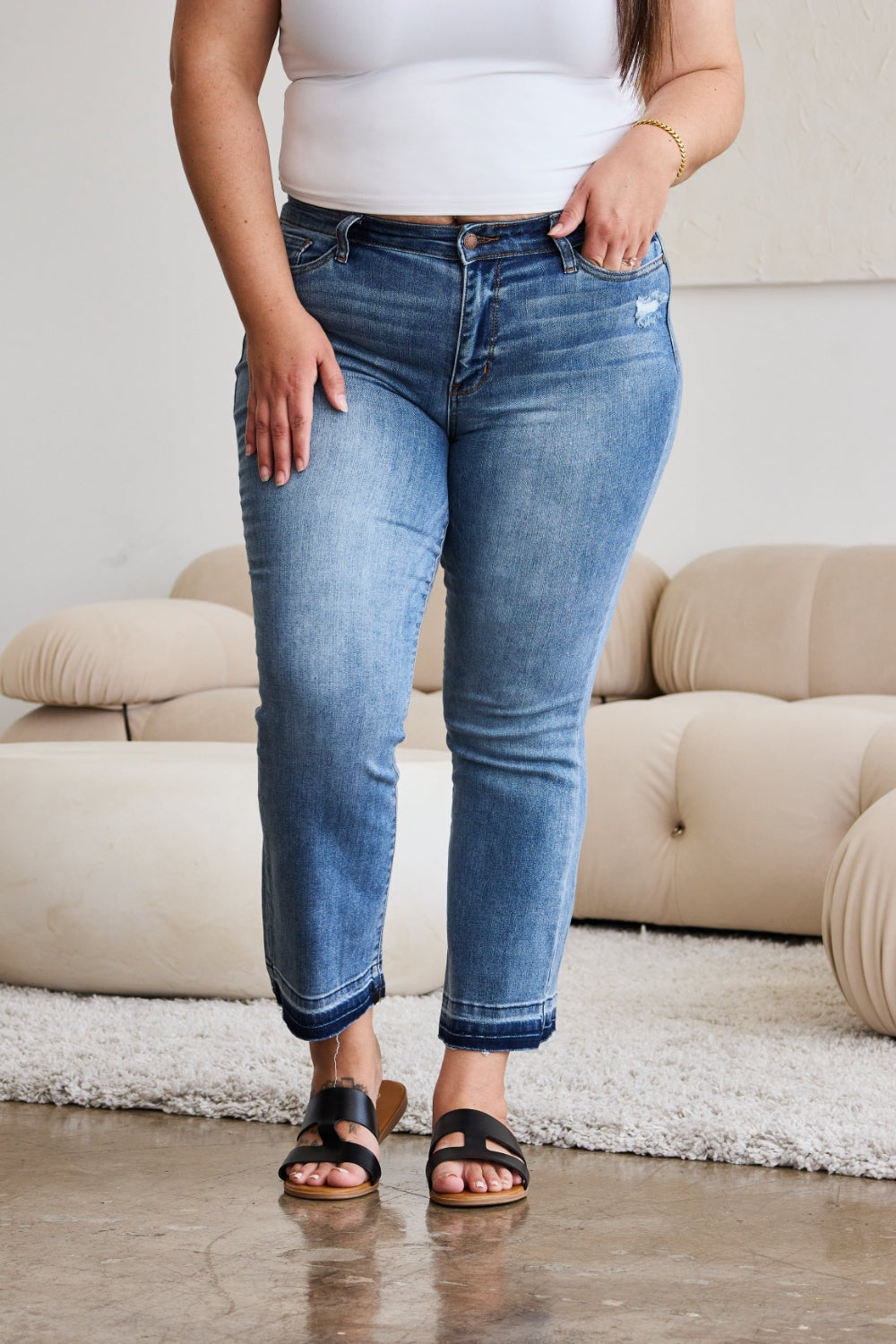 A person modeling Judy Blue Full Size Release Hem Cropped Bootcut Jeans and white heeled sandals stands in a room with light-colored furniture.