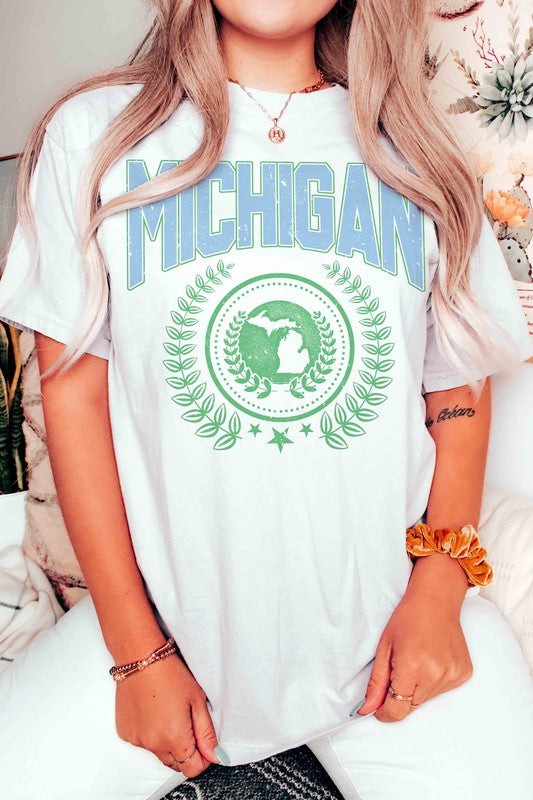 An individual dons a white MICHIGAN Graphic Tee, crafted from 100% cotton. The shirt prominently displays the word "Michigan" in blue, alongside an Earth graphic encircled by laurel leaves and topped with a star. Its unisex sizing guarantees a comfortable fit for all wearers.