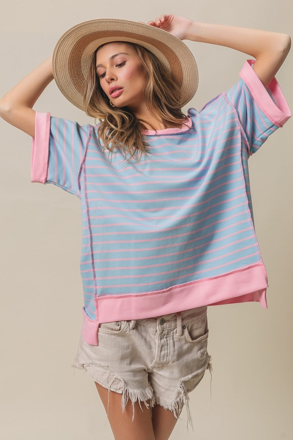 A person wearing the BiBi Exposed Seam Stripe Contrast T-Shirt in light blue and pink, paired with beige shorts and a wide-brimmed straw hat, stands against a plain background.