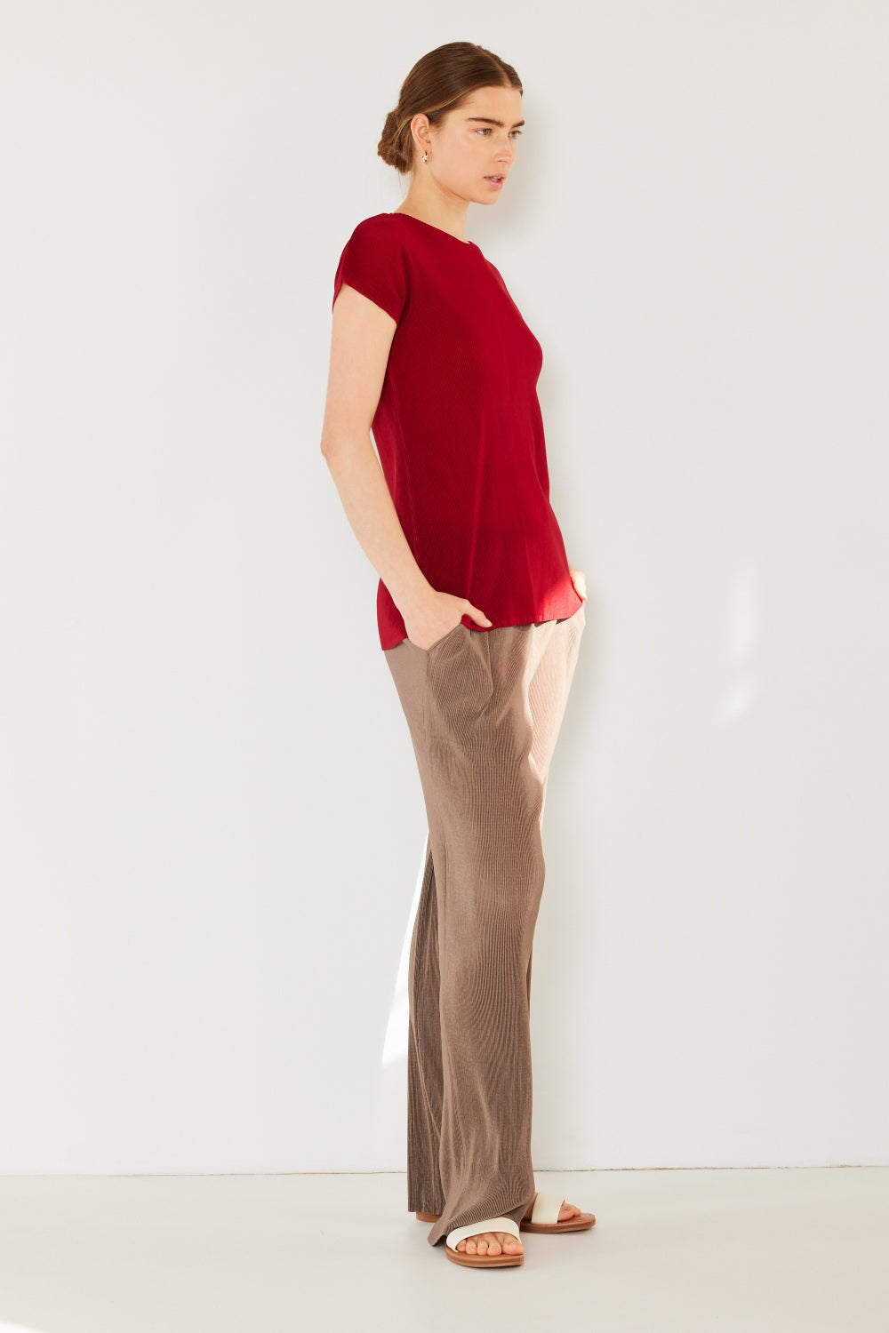 A person stands against a white wall, wearing a red short-sleeve shirt, Marina West Swim rib pleated elastic-waist wide-leg pants in beige, and white sandals. Their hands are in their pockets, and they sport a neutral expression.