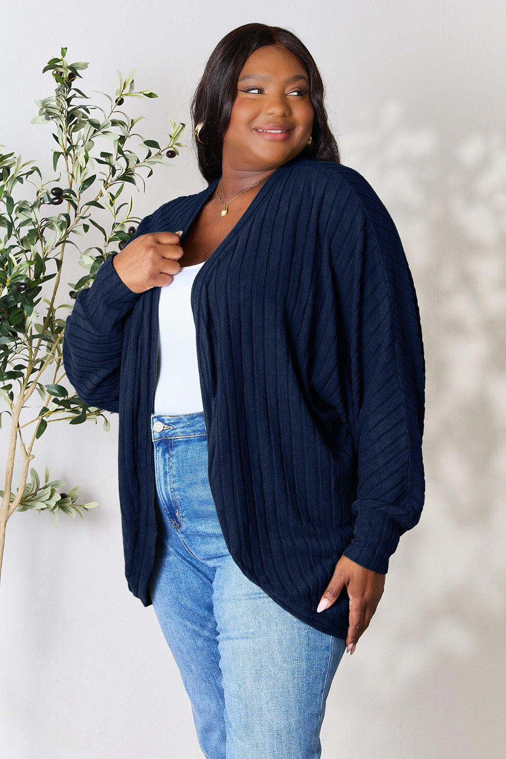 A person with wavy hair, wearing the Basic Bae Full Size Ribbed Cocoon Cardigan over a white top and beige pants, stands in front of a light-colored background with a leafy plant, showcasing an effortlessly basic style.