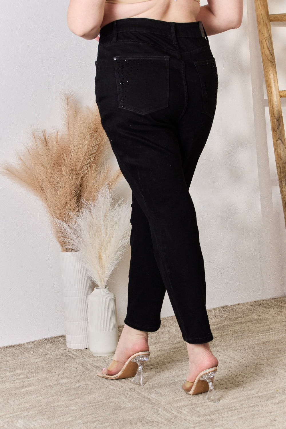 A person wearing slightly stretchy, black Judy Blue Full Size Rhinestone Embellished Slim Jeans and black shoes stands on a textured floor next to a white vase with brown and white ornamental grasses and a wooden ladder.