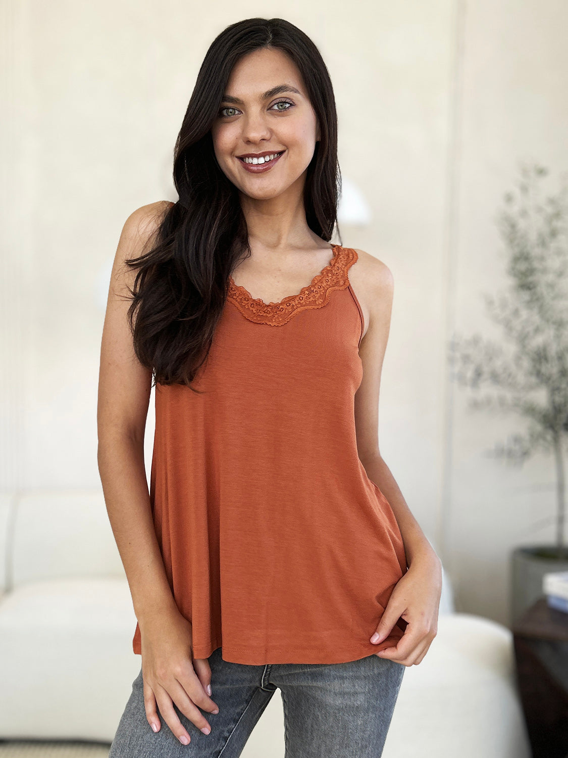 A woman with long hair wearing sunglasses, a slightly stretchy pink "Basic Bae Full Size Lace Detail V-Neck Cutout Cami," and basic style blue jeans stands against a concrete wall, looking to the side with one hand touching her upper chest.