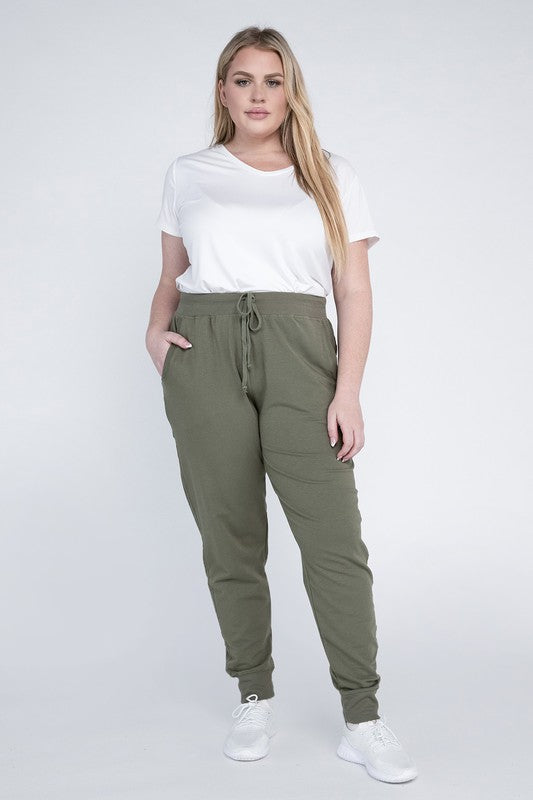 A person stands against a white background, wearing a white t-shirt, grey Plus-Size Jogger Pants with an adjustable waistband, and white sneakers. They have long blonde hair and are posing with one hand on their hip.