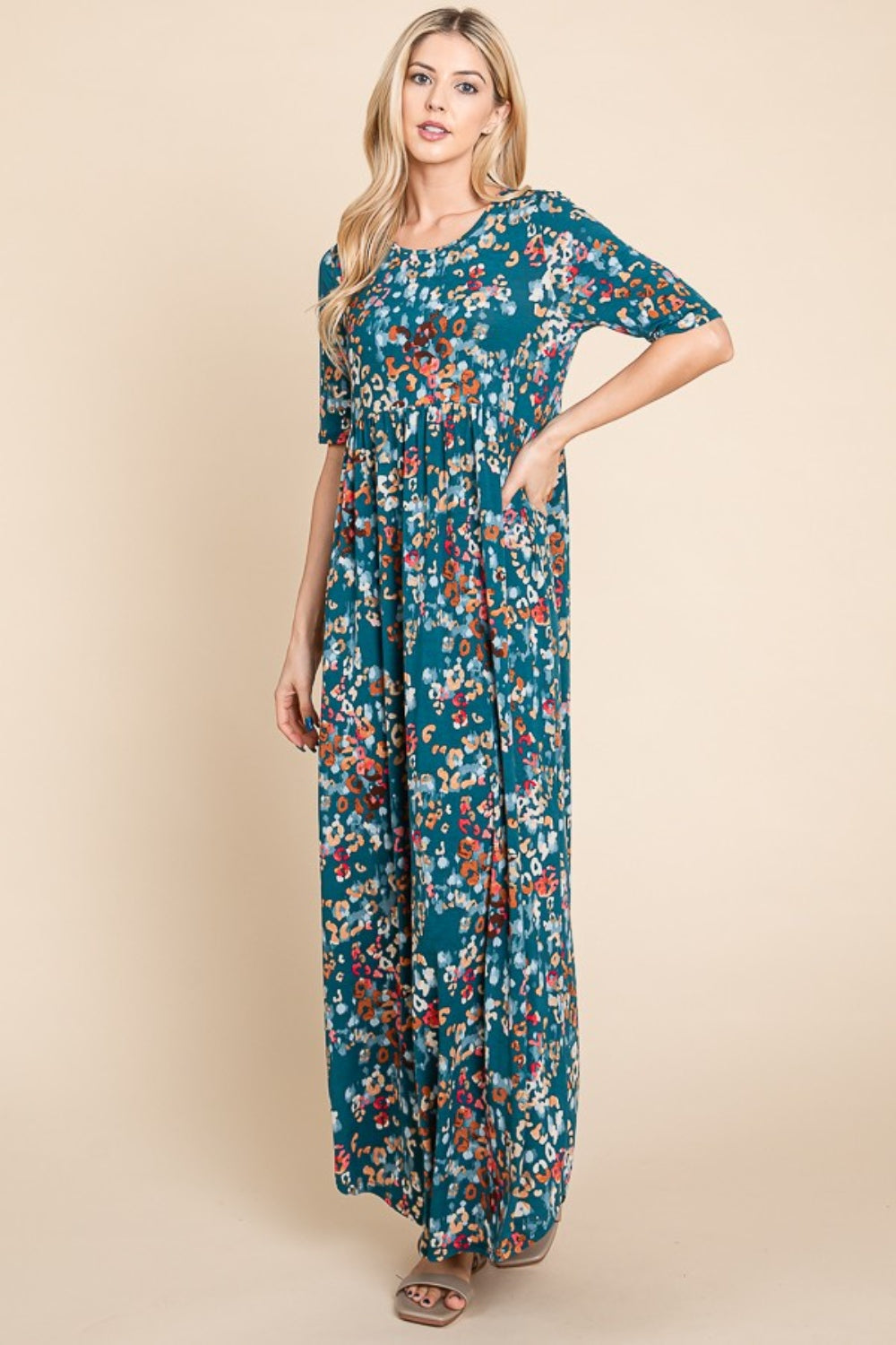 A person wearing a BOMBOM Printed Shirred Maxi Dress, featuring an eye-catching teal floral pattern and short sleeves, stands against a beige background.