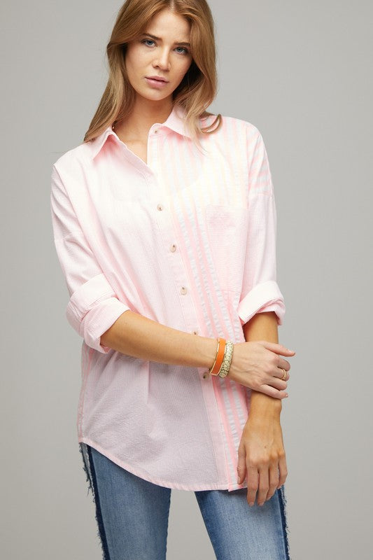 A person wearing a light pink Stripe Button Down Long Sleeve Shirt with rolled-up sleeves and jeans stands with their hands behind their head against a plain background.