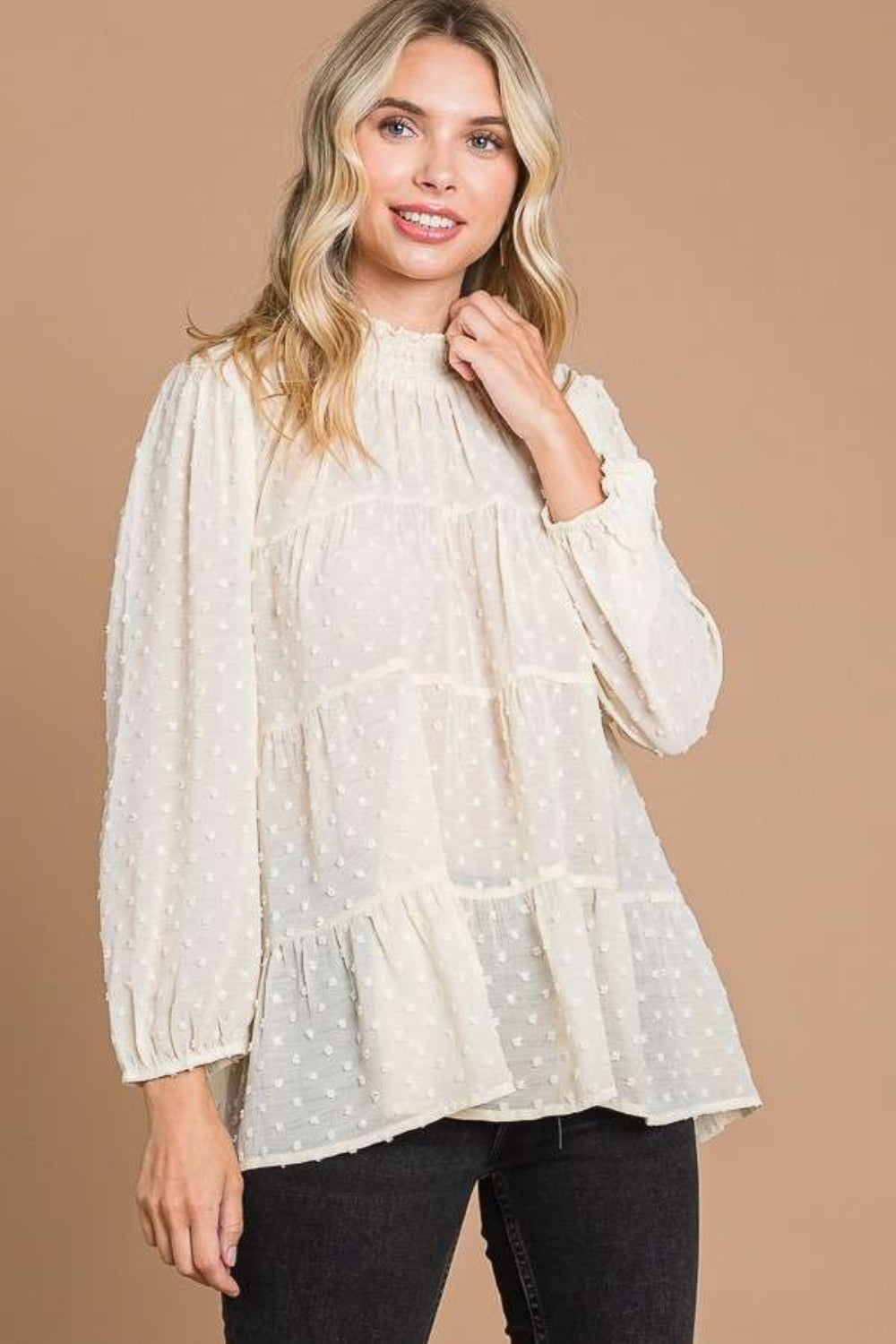 A woman with blonde hair stands against a plain brown background, exuding feminine fashion elegance in a loose, long-sleeved Culture Code Full Size Swiss Dot Smocked Mock Neck Blouse.