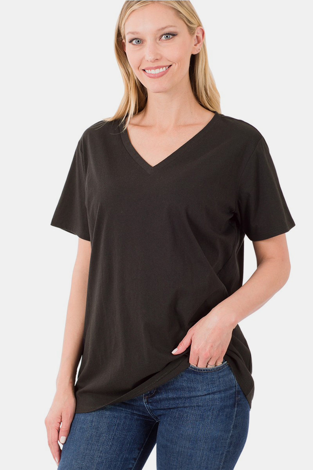 A blonde woman, wearing blue jeans and a black Zenana Full Size V-Neck Short Sleeve T-Shirt made of 100% cotton, stands against a plain white background, smiling and holding the hem of her T-shirt.