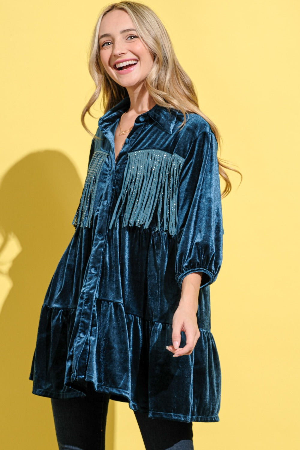 A woman in the And The Why Fringe Detailed Velvet Shirt Dress with a tailored fit and button-up front, wearing jeans, stands against a yellow background.