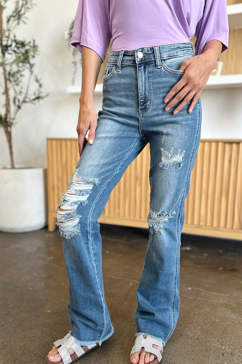 A person wearing Judy Blue Full Size Distressed Raw Hem Bootcut Jeans and a light purple top stands on a polished floor with a wooden cabinet in the background.