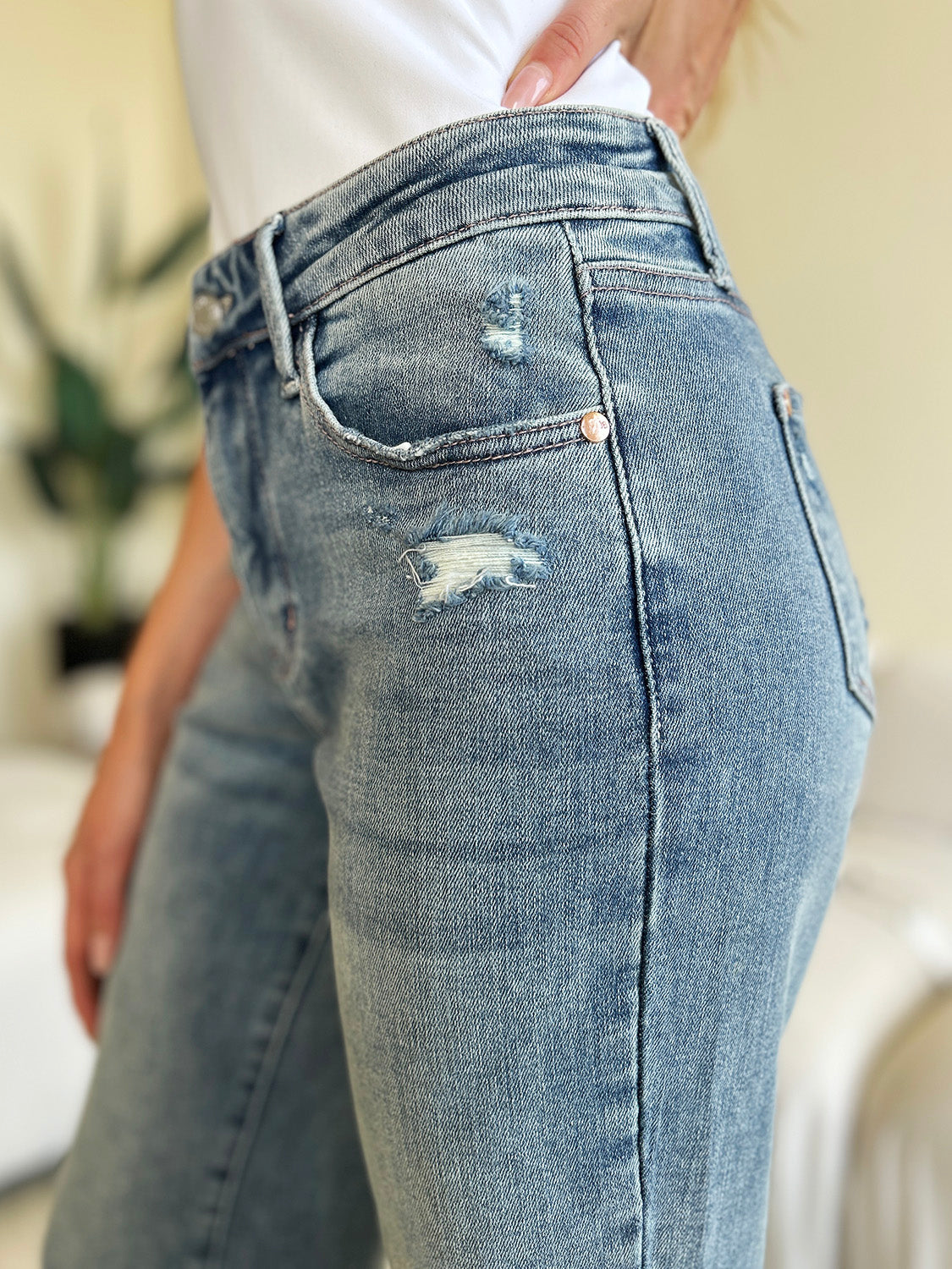 A person wearing Judy Blue Full Size Mid Rise Distressed Straight Jeans in light blue with a white top and white slip-on shoes stands in a room with a white couch and a plant.