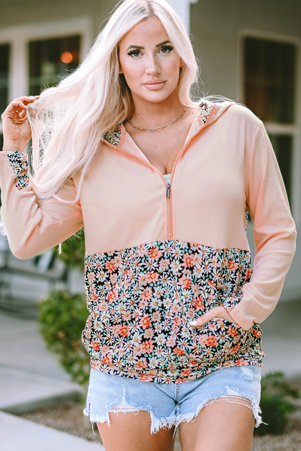 A person with long blonde hair wears a Pink Floral Patch Half Zip Kangaroo Pocket Hoodie and denim shorts, standing outdoors with their back facing the camera.