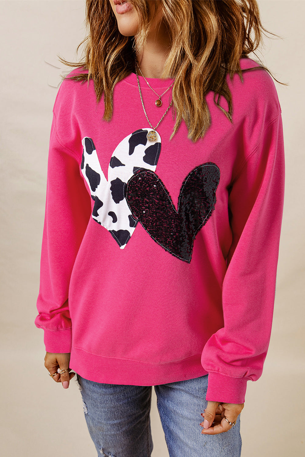 A person with long blonde hair, seen from the back, is wearing a Strawberry Pink Cow & Sequin Double Heart Patch Graphic Sweatshirt and light blue jeans.