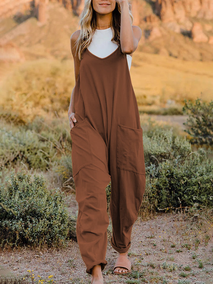 A woman stands outdoors wearing the Double Take Full Size Sleeveless V-Neck Pocketed Jumpsuit in a loose-fitting, imported brown design over a white top. Her surroundings include grass, bushes, and a rocky landscape in the background. The jumpsuit's polyester fabric ensures comfort and durability with every machine wash.