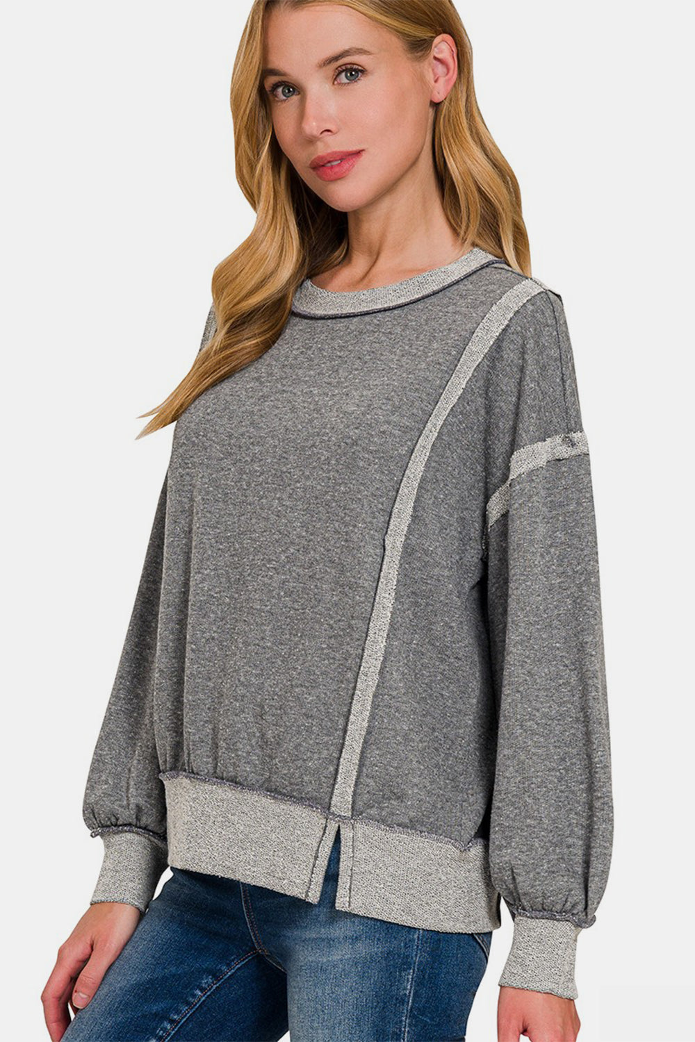 A woman with long blonde hair is seen in a neutral pose against a plain white background, wearing the grey Zenana Washed Exposed-Seam Sweatshirt featuring a crew neck and ribbed hem. Her casual wear is perfect for layering in any season.