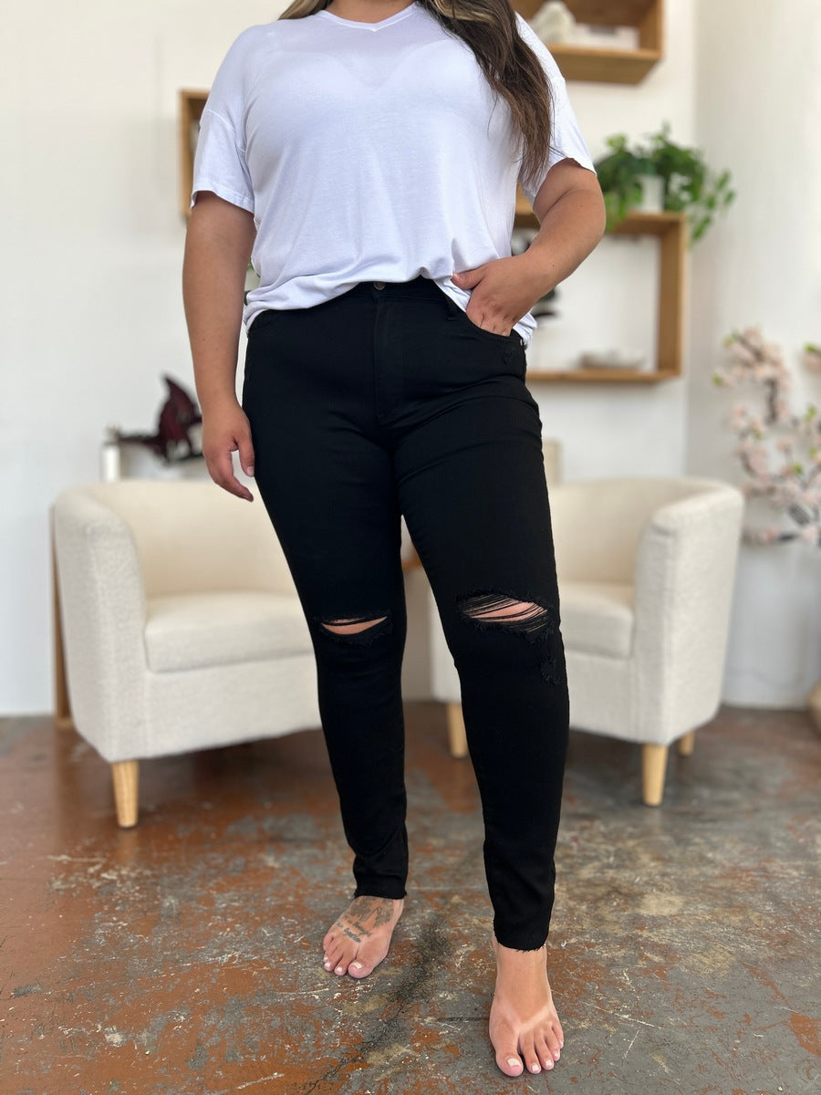 A person wearing a white T-shirt and Judy Blue Full Size Distressed Tummy Control High Waist Skinny Jeans poses barefoot in a room with white chairs and decorative plants in the background.