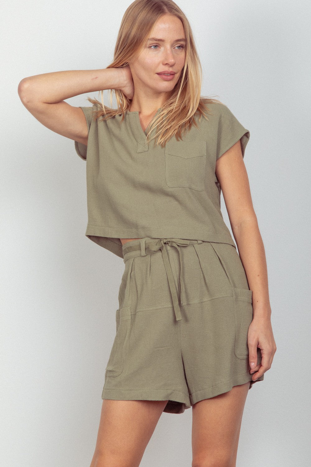 A person wearing the VERY J Woven Cropped Top & Waist Tie Shorts Set in khaki green, which includes a short-sleeved cropped top and matching waist tie shorts crafted from woven fabric, stands against a plain background.