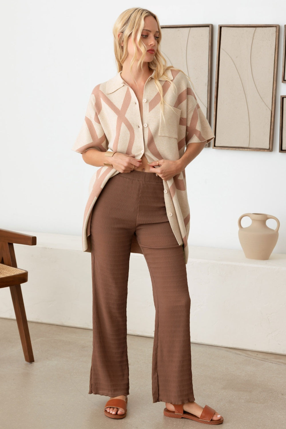 A woman with long blond hair wearing the Le Lis Abstract Collared Button Down Sweater Dress stands in a minimalist room with a wooden chair and ceramic vase—her versatile piece adds an abstract touch to the setting.