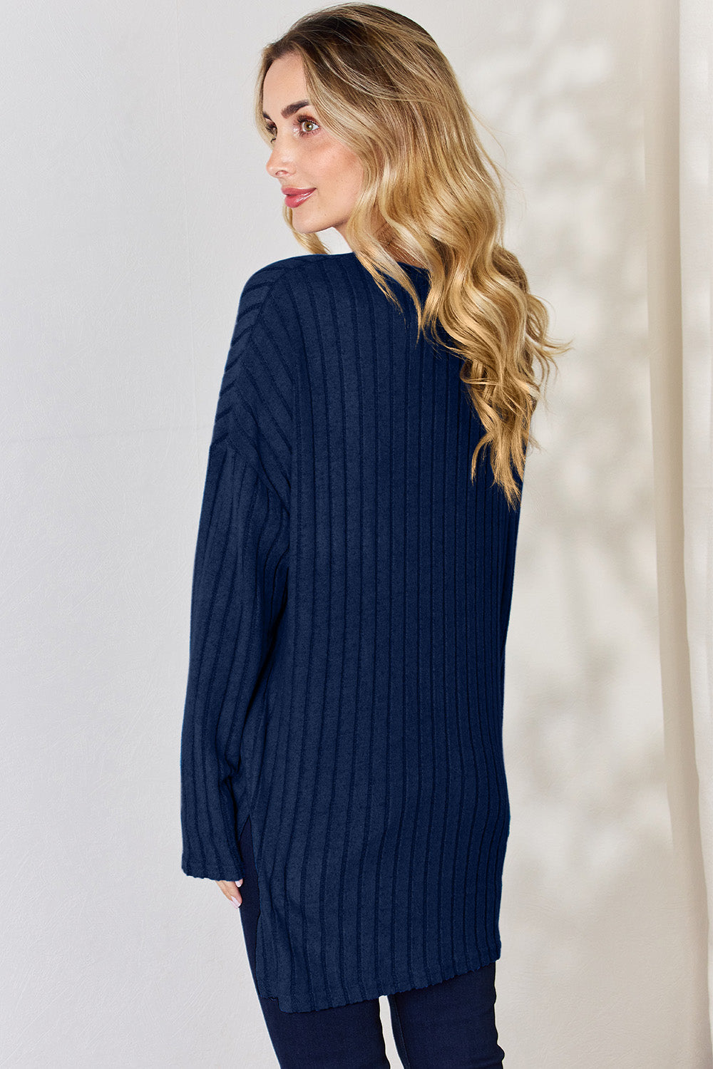 A person with long blonde hair is standing against a light background, wearing the Basic Bae Full Size Ribbed Half Button Long Sleeve High-Low T-Shirt in blue and dark pants. They are looking at the camera and smiling slightly, showcasing the chic high-low hem of their top.