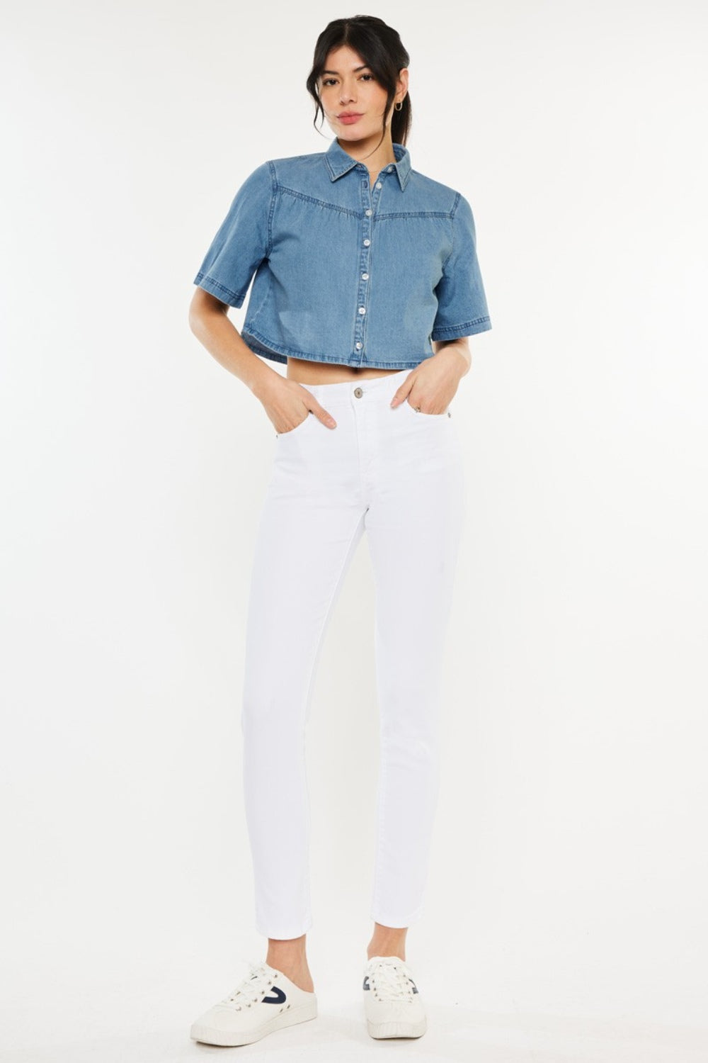 A person wearing a short-sleeve denim button-up shirt, Kancan High Rise Ankle Skinny Jeans in classic white, and white sneakers stands effortlessly against a plain background.