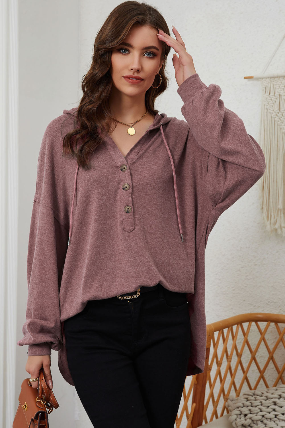 A woman stands facing away, wearing a stylish Buttoned High and Low Hem Hoodie in mauve made of high-quality fabric and black pants. She is in a room with light curtains and a wicker chair.