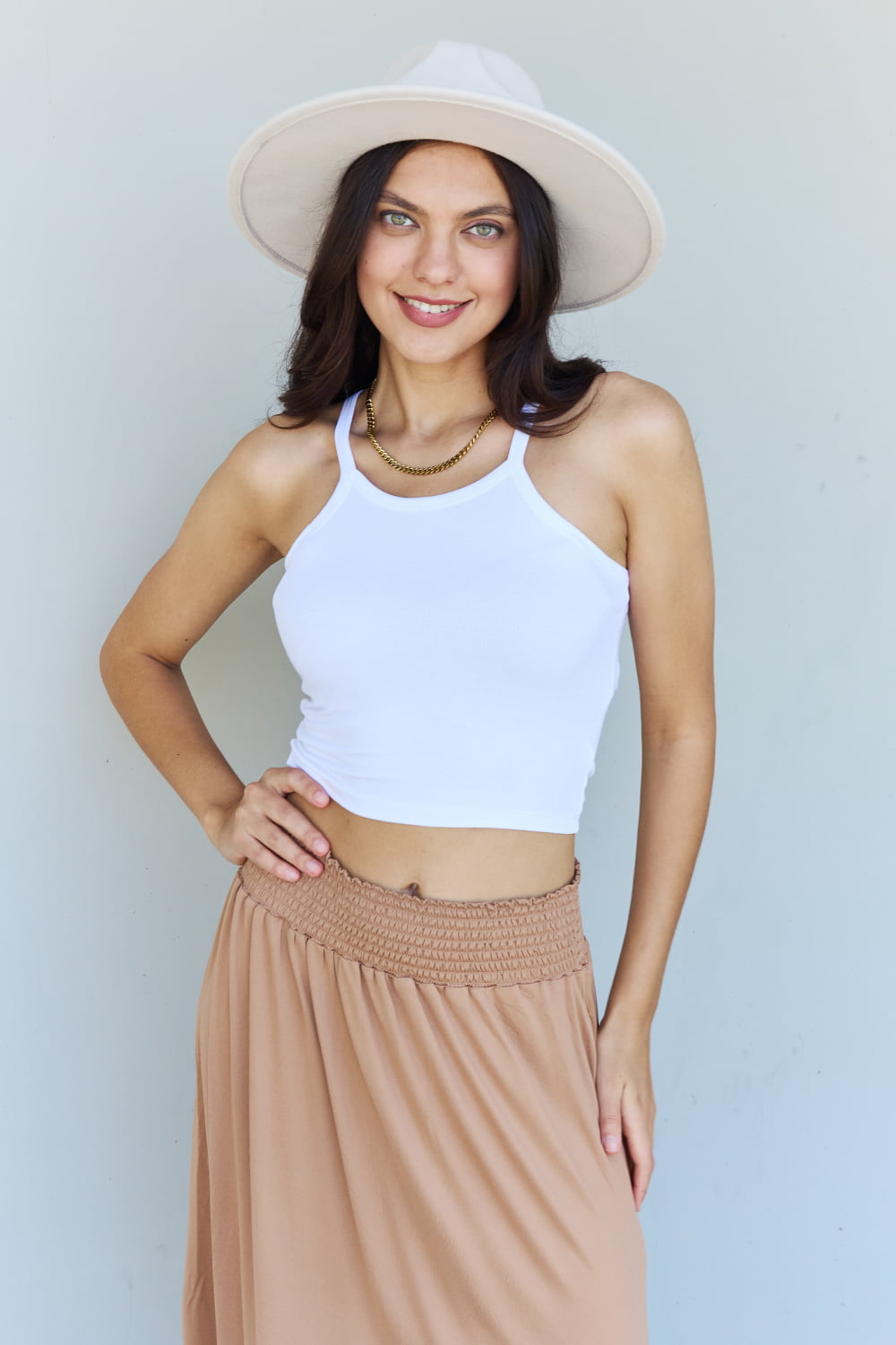 A person stands against a wall, wearing a white sleeveless top, the Doublju Comfort Princess Full Size High Waist Scoop Hem Maxi Skirt in Tan, and blue platform sandals. The right hand rests on the side of the skirt.