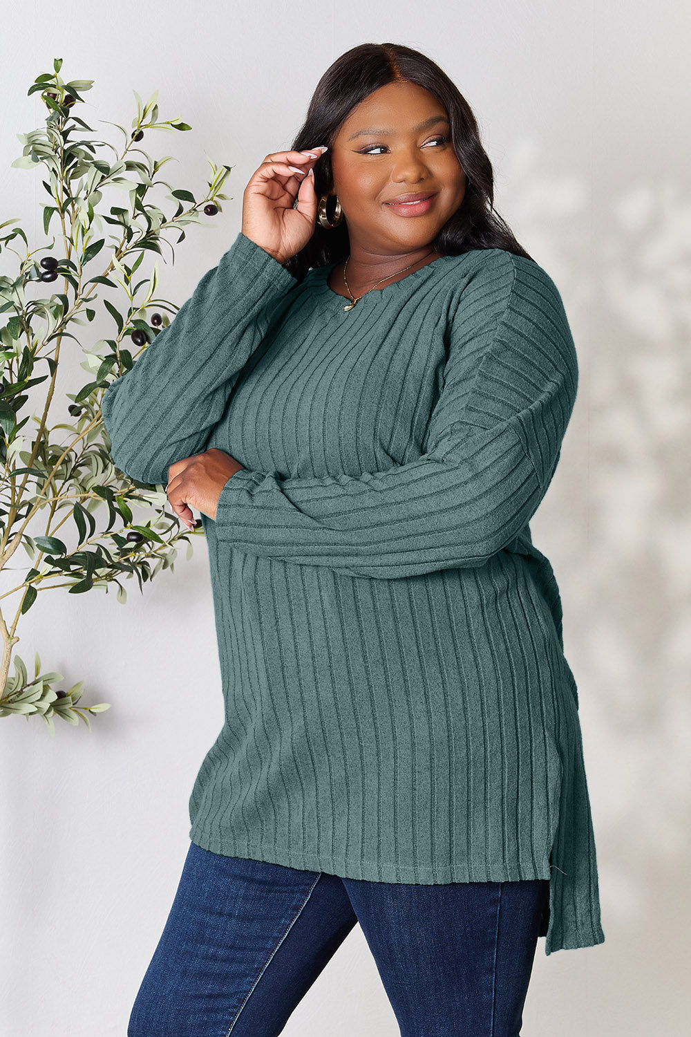 A person with shoulder-length hair smiles while wearing a beige ribbed sweater called the Basic Bae Full Size Ribbed Round Neck Long Sleeve Slit Top, along with matching beige pants, showcasing a basic style. They stand next to a green plant.