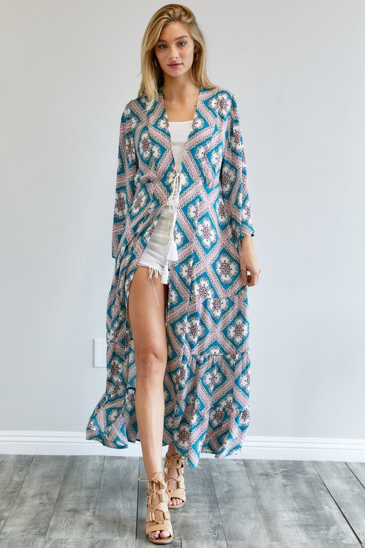 A person wearing a Printed Long Sleeve Loose Kimono with a bohemian blue and pink print over a white outfit stands in a room with a gray floor and white wall.