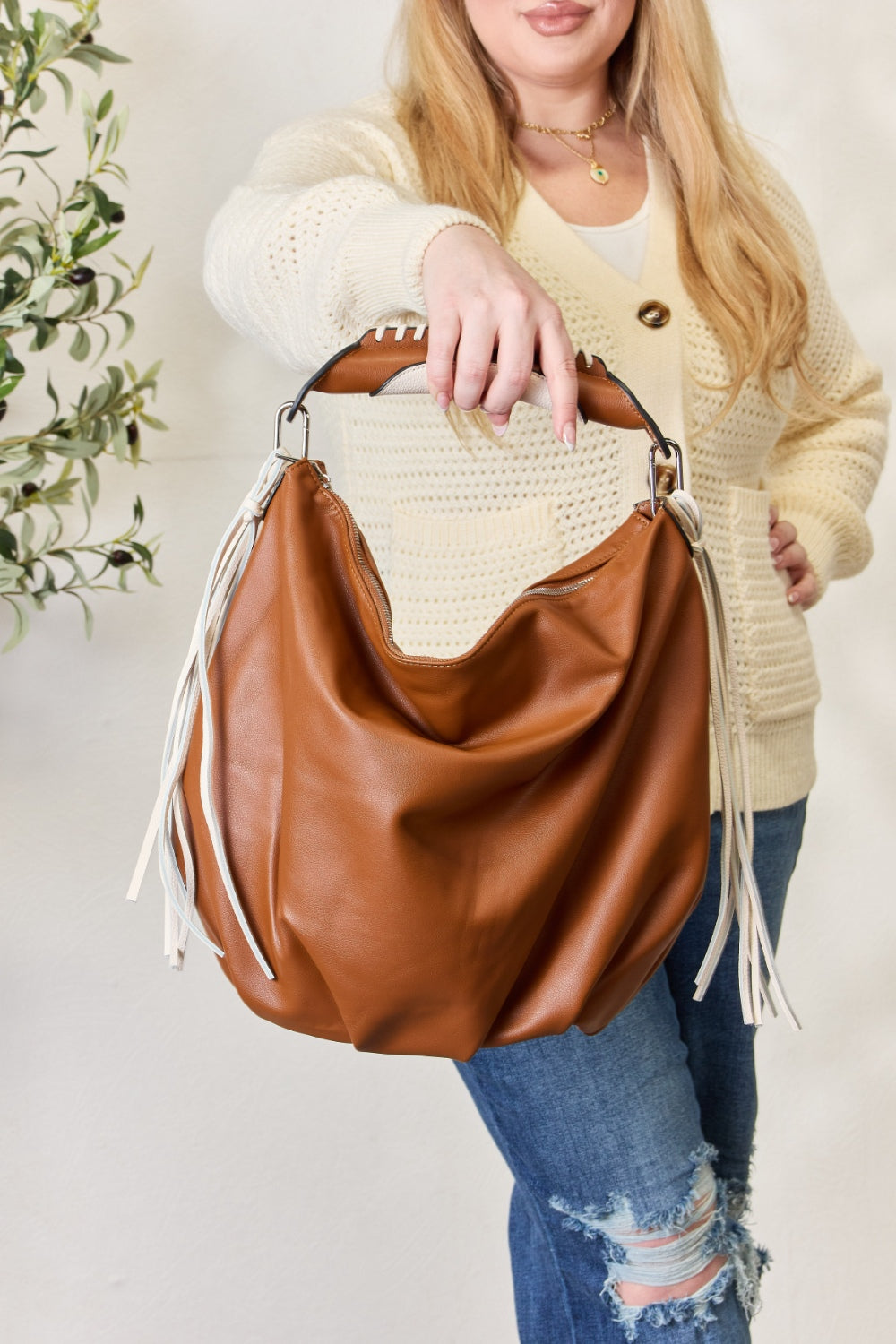 Individual holding a SHOMICO Fringe Detail Contrast Handbag, featuring cream and brown accents, paired with a knitted cream sweater, standing against a light background. The handbag exudes Bohemian flair with its relaxed style.