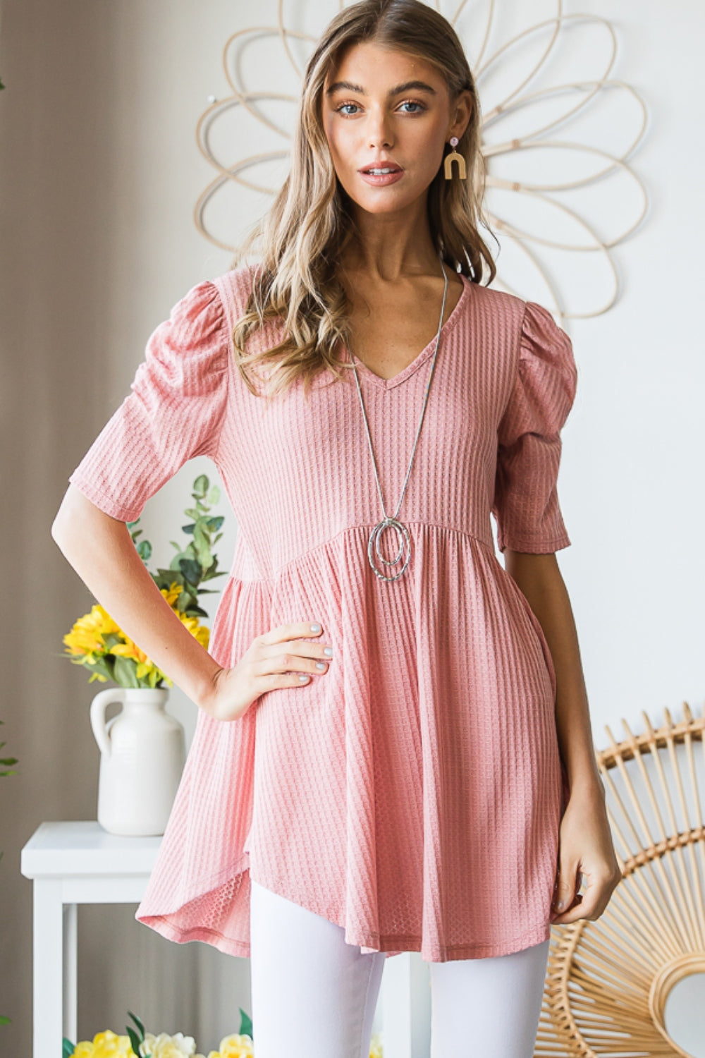 A woman in a Heimish Full Size Waffle Knit V-Neck Babydoll Top with short-sleeves and a long necklace, standing by a window with a neutral expression; the perfect cozy wardrobe addition.