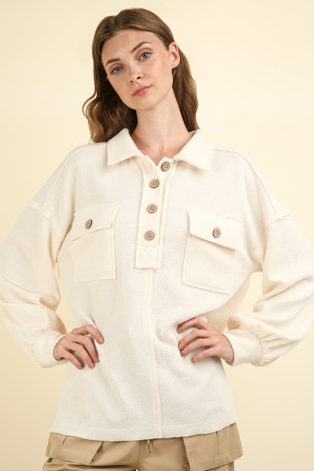 A woman with long hair stands against a neutral background, wearing an oversized VERY J Collared Half Button Knit Top with large pockets in cream.