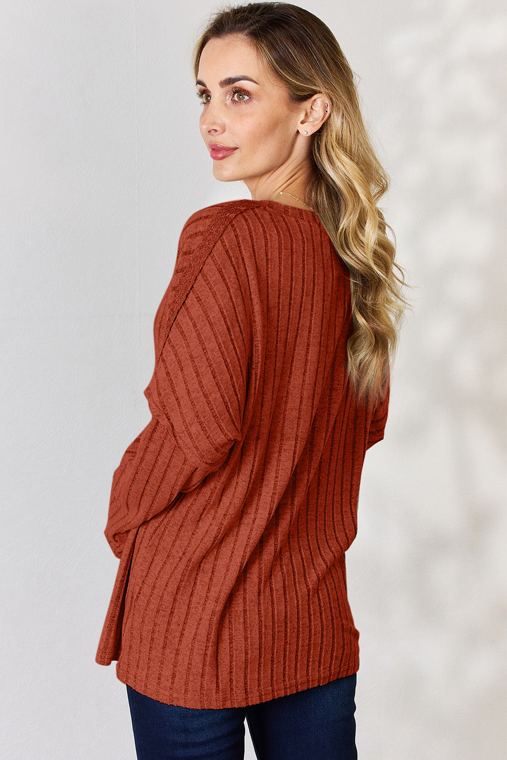 A woman with long, wavy blonde hair wears a slightly stretchy, textured Basic Bae Full Size Ribbed Half Button Long Sleeve T-Shirt in pink, standing against a neutral backdrop.