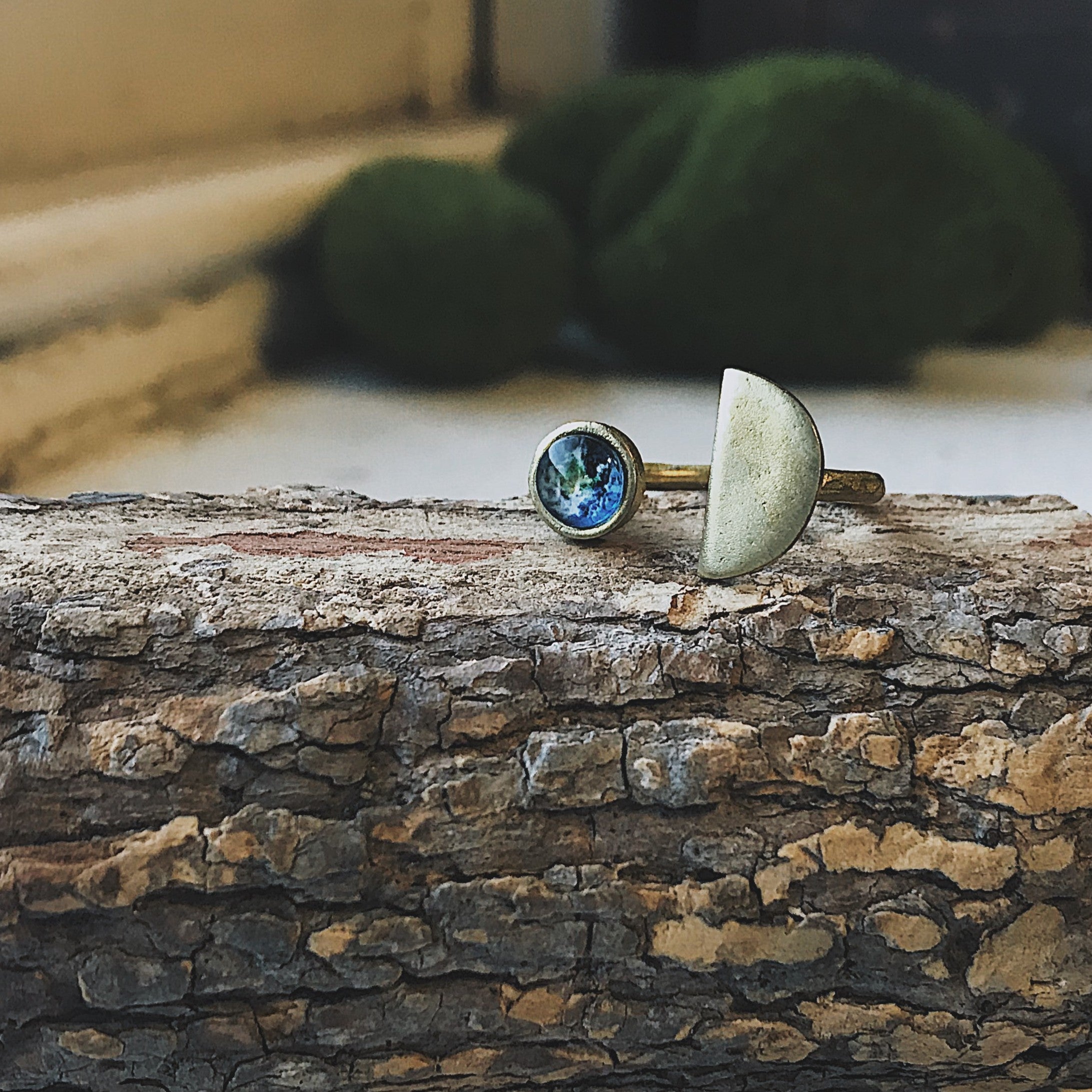 A hand displays two exquisite handcrafted jewelry pieces: one, the Earth Rise Ring, has a small Earth image reminiscent of Apollo 8, and the other showcases a simple, elegant half-circle shape.