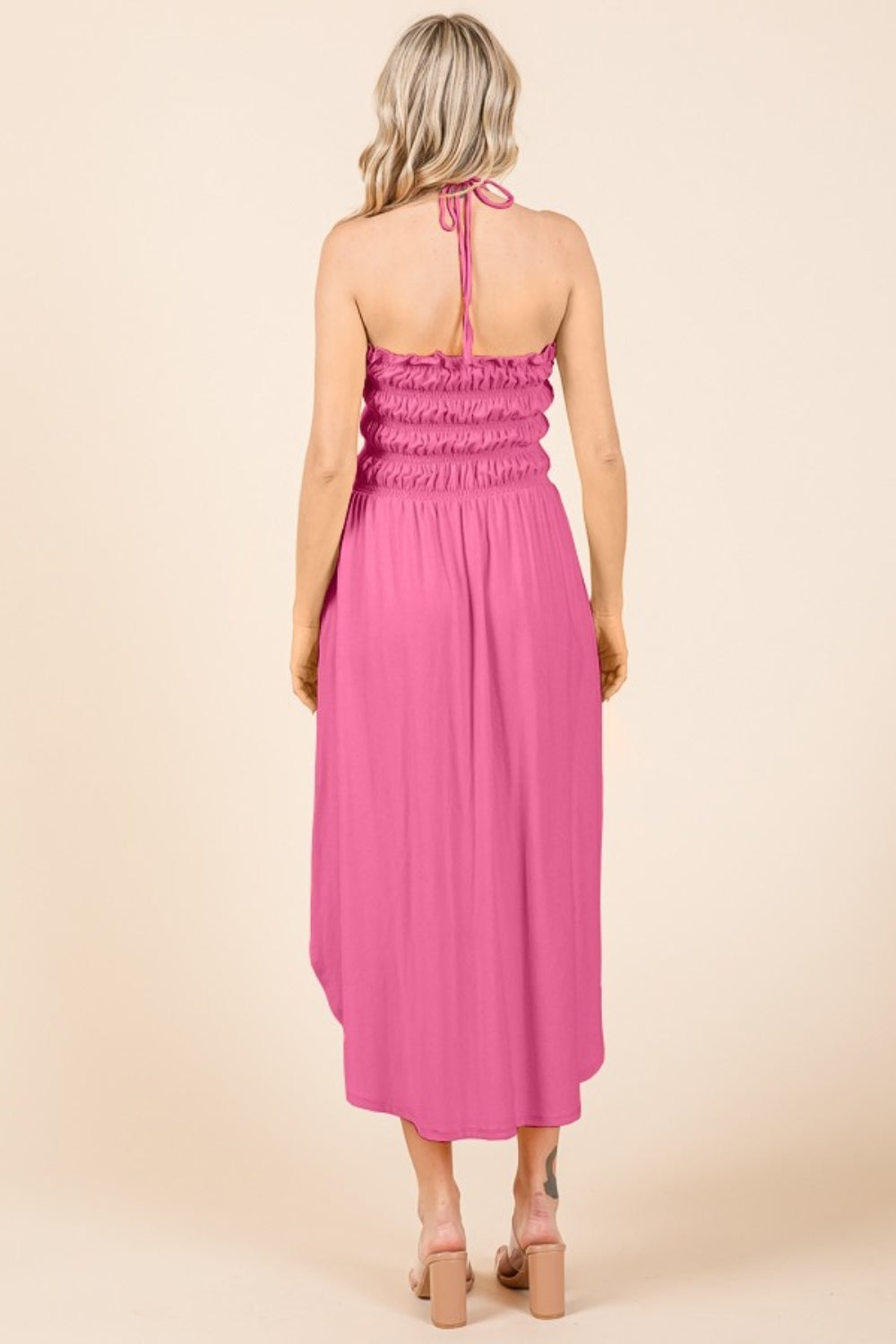 Someone wearing a Culture Code Tie Back Shirring Dress with pockets, featuring a sleeveless and smocked pink halter style, standing against a beige background.